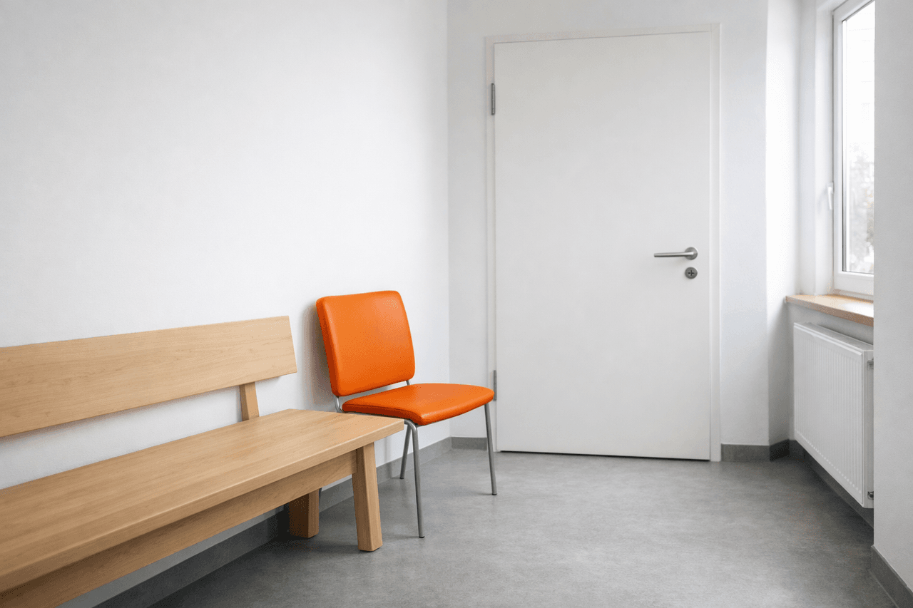 Heller Flur in einer Praxis mit Holzbank, einem orangen Stuhl und einer geschlossenen weißen Tür, rechts ein Fenster