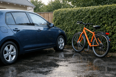Dunkelblaues Auto auf nasser Einfahrt, daneben ein oranges Fahrrad vor einer Hecke